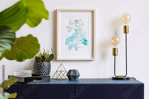 White interior of floral poster mock up with vertical wooden frame, table lamp, , cacti, gold pyramid and tropical flowers  on the white wall background. Concept with navy blue shelf.