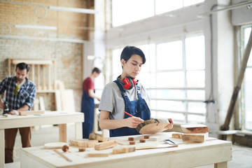 Wall Mural - Serious busy masculine girl with ear protectors on neck standing at table and drawing on slice of wood while making childrens toys