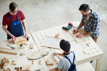 Directly above view of busy concentrated carpenters standing at table and making wooden detail for interior design, men drawing outline