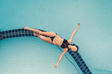 Fototapeta Tęcza - Aerial Top View of Beautiful sexy girl in a black bikini lying on the edge of the swimming pool with transparent turquoise water. Relax, sunbathing, fun, holidays, resort on tropical island