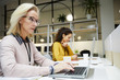 © Seventyfour - Busy staff in office: serious thoughtful blond-haired mature lady in glasses sitting at table and typing on laptop while preparing report