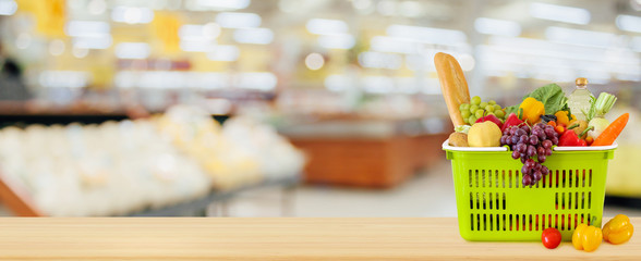 Wall Mural - Shopping basket filled with fruits and vegetables on wood table with supermarket grocery store blurred defocused background with bokeh light