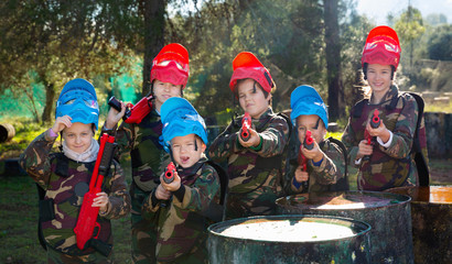 Wall Mural - Friendly group of children paintball players in camouflage posing with guns on paintball playing field outdoors