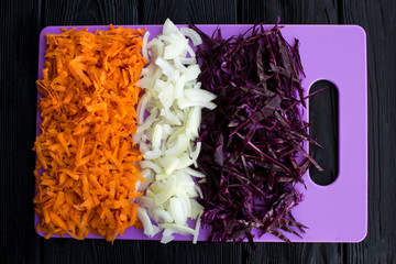 Wall Mural - Chopped fresh blue cabbage,onion and carrot on the violet cutting board on the black wooden background.Top view.