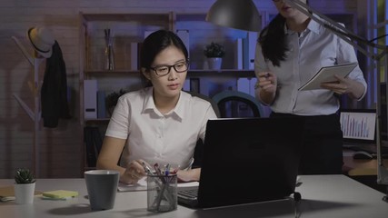 Wall Mural - young asian female colleague asking problem discussing working together in dark office at night. businesswomen in smart casual using calculator laptop computer. girl worker walk find document file