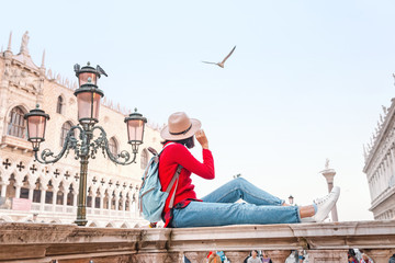 Wall Mural - Young happy asian girl traveler with hat and backpack sitting on a parapet on San Marco square in Venice. Vacation and holidays in Italy and Europe concept
