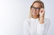 Successful stylish and elegant young lady in glasses and sweater looking delighted with happy pleased smile at upper left corner checking eyewear thinking or daydreaming over white wall