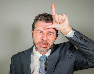 unshaven sad and ashamed businessman crying doing loser sign with hand and fingers on his forehead with funny depressed expression in unsuccessful career concept