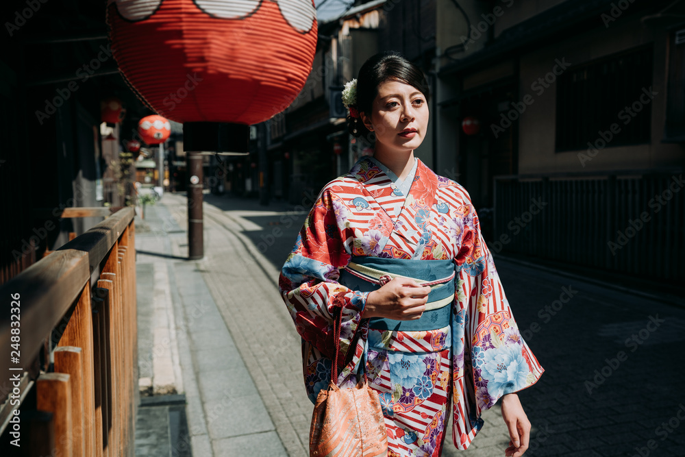 Woman in traditional Japanese kimono walking peacefully through historic Kyoto street with traditional wooden architecture and red lanterns