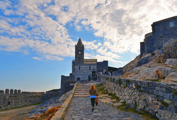 Wall Mural - San Pietro church, Lord Byron Natural park with young woman in Portovenere village on stone cliff rock and blue sky, view from Doria castle, Riviera di Levante, La Spezia, Liguria, Italy 