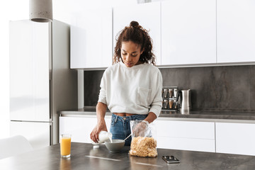 Wall Mural - Image of happy african american woman making breakfast with corn flakes and milk, in stylish kitchen
