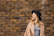 © LUMINA IMAGES - Portrait of beautiful smiling Caucasian woman standing by the brick wall wearing camel coat and a hat.