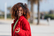 © 2Design - Front view of a young smiling curly afro woman standing outdoors while smiling and listening music by earphones in a sunny day