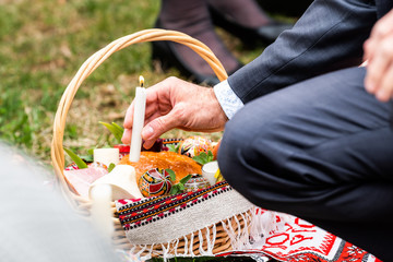 Ukrainian Orthodox Easter blessing wicker straw baskets with senior man hand placing candle on grass ground outside at church with flame