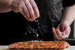 © spaskov - Raw piece of meat, beef ribs. The hand of a male chef puts salt and spices on a dark background.