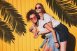 © VlaDee - Candid shot of two lovely hipster women in glasses and denim clothes standing under the shadow of palm branches on a sunny summer day.