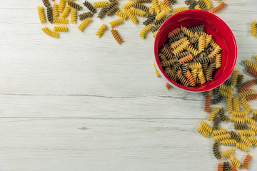 Wall Mural - Raw uncooked three-colored Fusilli  in a red bowl on white wood table.