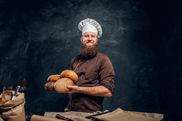 A bearded baker wearing a uniform holding fresh bread, standing next to a table, decorated with delicious bread loaves, baguettes and muffins in a dark studio