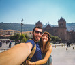 handsome happy tourist couple taking selfie in Plaza de Armas with Inca fountain, Cathedral and Compania de Jesus Church, city of Cusco, Peru