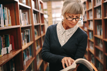 Wall Mural - Senior woman looking at a book standing in library