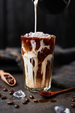 Ice Coffee In A Tall Glass With Cream Poured Over And Coffee Beans On Dark Concrete Table Over Black Wooden Background. Cold Summer Drink. Copy Space For Text. Selective Focus.