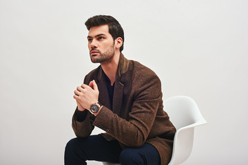 Searching for solution. Stylish dark-haired man sitting on a chair and looking at camera isolated over white background