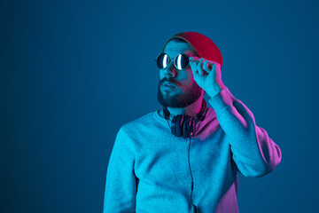 Enjoying his favorite music. Happy young stylish man in hat and sunglasses with headphones listening and smiling while standing against blue neon background