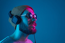 Enjoying His Favorite Music. Happy Young Stylish Man In Hat And Sunglasses With Headphones Listening And Smiling While Standing Against Blue Neon Background