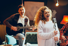 Band Practice For The Show. Woman With Curly Hair Holding Microphone And Singing While Man In Background Playing Acoustic Guitar. Home Studio Interior.