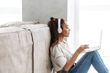 Wall Mural - Photo of dreamy african american woman wearing headphones using laptop, while sitting on floor in bright living room
