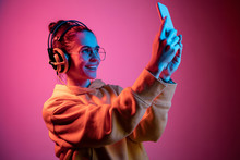 Fashion Pretty Woman With Headphones Listening To Music And Making Selfie Photo Over Red Neon Background At Studio.