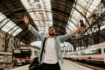 Attractive positive businessman feeling himself happy after long business travel trip.Casual professional entrepreneur using smartphone at hall of train station