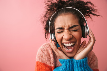 Canvas Print - Emotional beautiful young african woman posing isolated over pink wall background listening music with headphones.