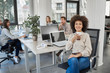 © nenadaksic - Smiling mixed race CEO sitting at office and drinking coffee while looking at camera. In background employees working.
