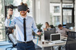 Employee in formal wear trying out VR technology in office. In background other employees working.