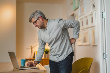 middle aged gray haired man having backache while working laptop at his home