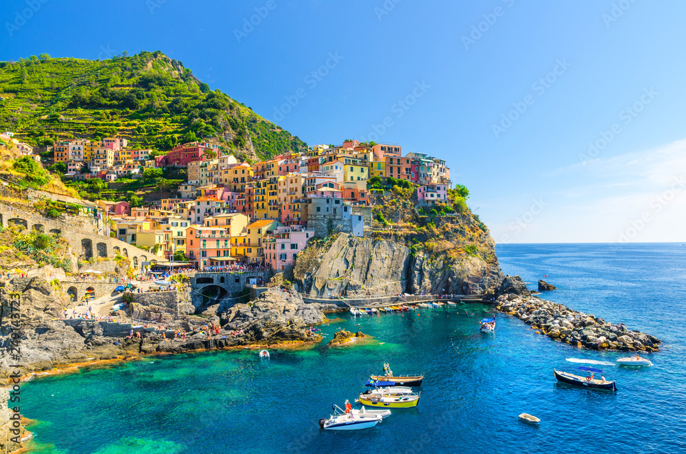 Manarola traditional typical Italian village in National park Cinque Terre, colorful multicolored buildings houses on rock cliff, fishing boats on water, blue sky background, La Spezia, Liguria, Italy - obrazy, fototapety, plakaty 