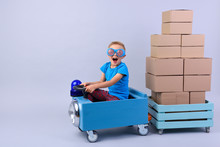Smiling Boy Driving Blue, Fanny Car And Wagon With Small Boxes.Happy Child With Paper Boxes On Grey Background. Studio Shot. Delivery, Shipping,  Transport And Supplier Concept. 