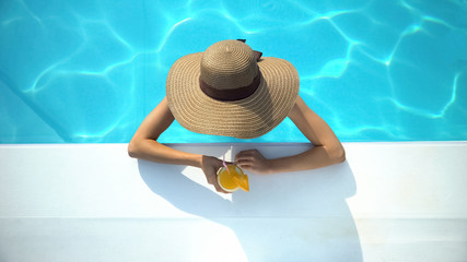 Young woman in elegant hat swimming in pool and enjoying delicious cocktail