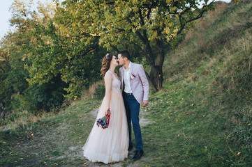  Autumn portrait of newlyweds in love, on a background of green trees on nature. Wedding photography. Stylish groom in a plaid jacket hugging a pretty bride in a beige dress with a crown.