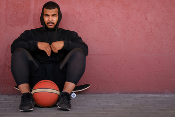 Wall Mural - African-American man wearing a black hoodie sitting on a skateboard and holding a basketball while leaning on a wall outside