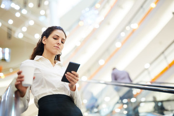 Wall Mural - Young elegant businesswoman with drink scrolling in smartphone while moving on escalator