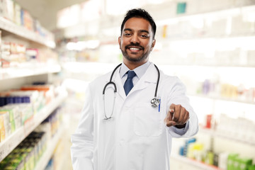 medicine, pharmacy and healthcare concept - smiling indian male doctor or pharmacist in white coat with stethoscope pointing finger to you over drugstore background