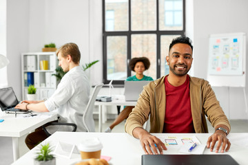 business and people concept - happy smiling indian male creative worker with laptop sitting at office table