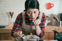 Young Asian Female Cook In Apron Melting Chocolate In Hot Pot On Stove Using Finger Tasting Flavor Looking Aside. Elegant Chinese Woman Make Sweet Dessert For Boyfriend On Valentine Day As Gift.