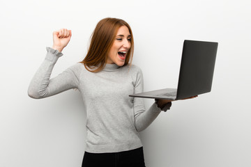 Redhead girl over white wall with laptop and celebrating a victory