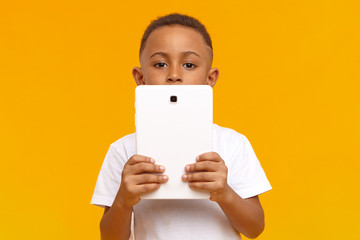 Wall Mural - Picture of smart dark skinned schoolboy holding generic white digital tablet, doing homework, looking for information on internet. Cute curious African American male child using touch pad pc