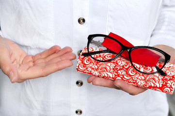 Canvas Print - Ophthalmologist holding eyeglasses for a try out. Optometrist hands holding new black plastic eyeglasses red microfiber on white-red case, at optician shop.Close-up view focused on glasses. 