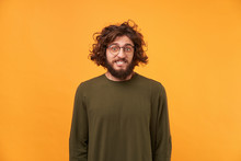 A Man With A Beard In Glasses And Curly Dark Hair Looking At The Camera, Bite Lip, Confused Bewildered As If Made Mistake, Feel Guilty Isolated On A Yellow Background Dressed In Ordinary Clothes.