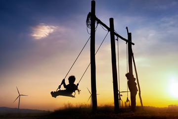 Silhouettes of happiness life of child to playing swing in turbine wind park renewable on sunset. Family Responsibility, Corporate Social Responsibility and sustainable energy concept.